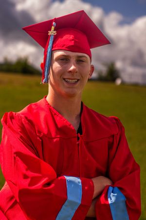 A graduate in bright red cap and gown stands outdoors on a sunny day with puffy clouds in the background.