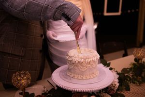 A white wedding cake with delicate piping and decorative details sits on a pink-lit table at a reception.