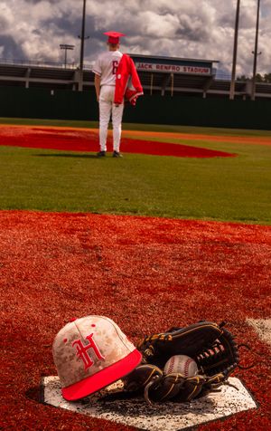 Baseball equipment including a red cap and glove rests on home plate on a red clay baseball field.