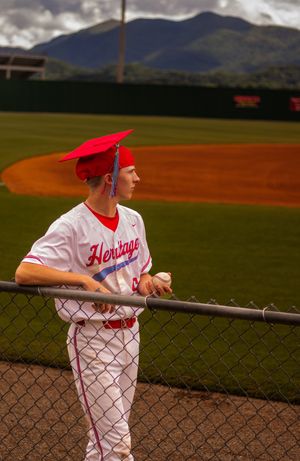 Baseball player in white and red uniform leans against chain link fence during game with mountains in background.