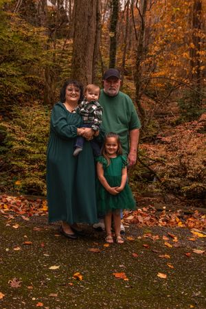 A family in matching green outfits poses together on fallen autumn leaves in a forest setting.