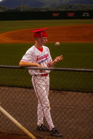 Baseball player in white uniform and red graduation cap tosses a ball while standing by chain link fence on baseball field.