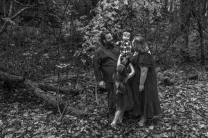 A group of people sit among autumn leaves in a wooded area during a casual outdoor photoshoot.