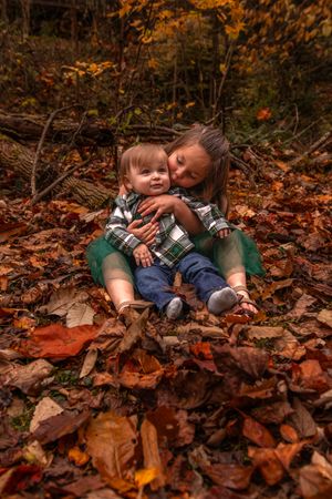 Two children sitting together among colorful autumn leaves in the forest during fall season.