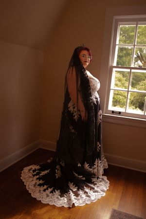 A person wearing an elegant black lace dress with white trim stands in dramatic lighting by a window.