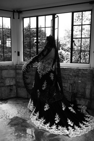 A dark figure in an elegant black dress with white lace trim stands in an abandoned room with large windows.