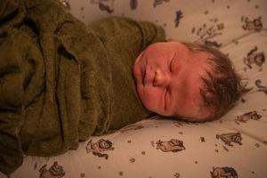 A sleeping baby rests on patterned bedding while wearing a brown sweater.