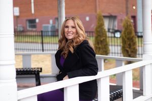 Professional wearing a black coat smiles while leaning against white porch railing on an autumn day.