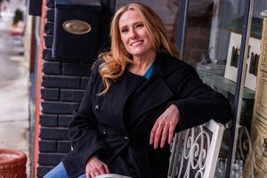A person in a black coat smiles while standing outside a storefront window display.