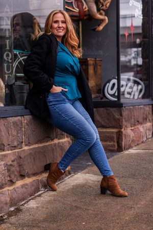 Woman in blue top and jeans poses casually against brick wall outside storefront in brown boots.