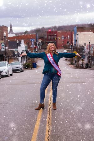 Person in blue jacket strikes joyful pose in falling snow on small town street.