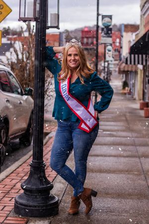 A person wearing a sash and jeans poses playfully next to a black lamppost on a downtown sidewalk during winter.
