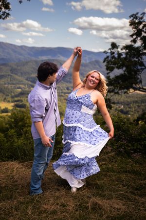 A couple dances together on a scenic mountaintop overlook with rolling hills and mountains in the background.