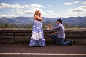 A romantic proposal moment on a scenic mountain overlook with blue skies and mountains in the background.