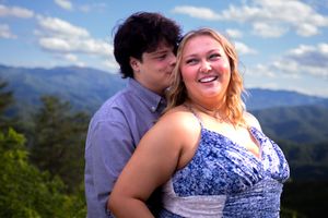 A couple in formal attire embraces while standing on a mountain overlook with scenic views on a sunny day.