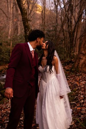 A romantic couple in wedding attire shares intimate moments in a wooded autumn setting with fallen leaves.