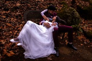 A couple in wedding attire pose dramatically on autumn leaves in a forest setting.