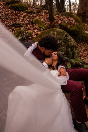 A romantic wedding couple embraces in a forest setting with autumn leaves and a flowing white dress.