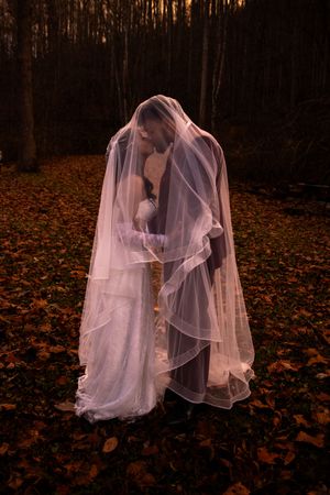 Ethereal figure draped in flowing white veil stands among fallen autumn leaves in dark moody forest at dusk.