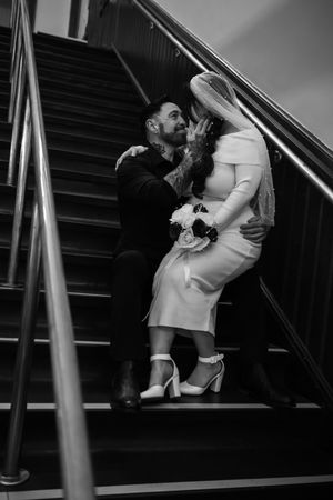 An intimate black and white wedding photo captures a tender moment between newlyweds on a staircase.