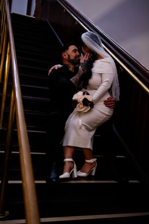 A romantic moment on dark stairs as a couple embraces, with the bride wearing a chic white dress and holding a bouquet.