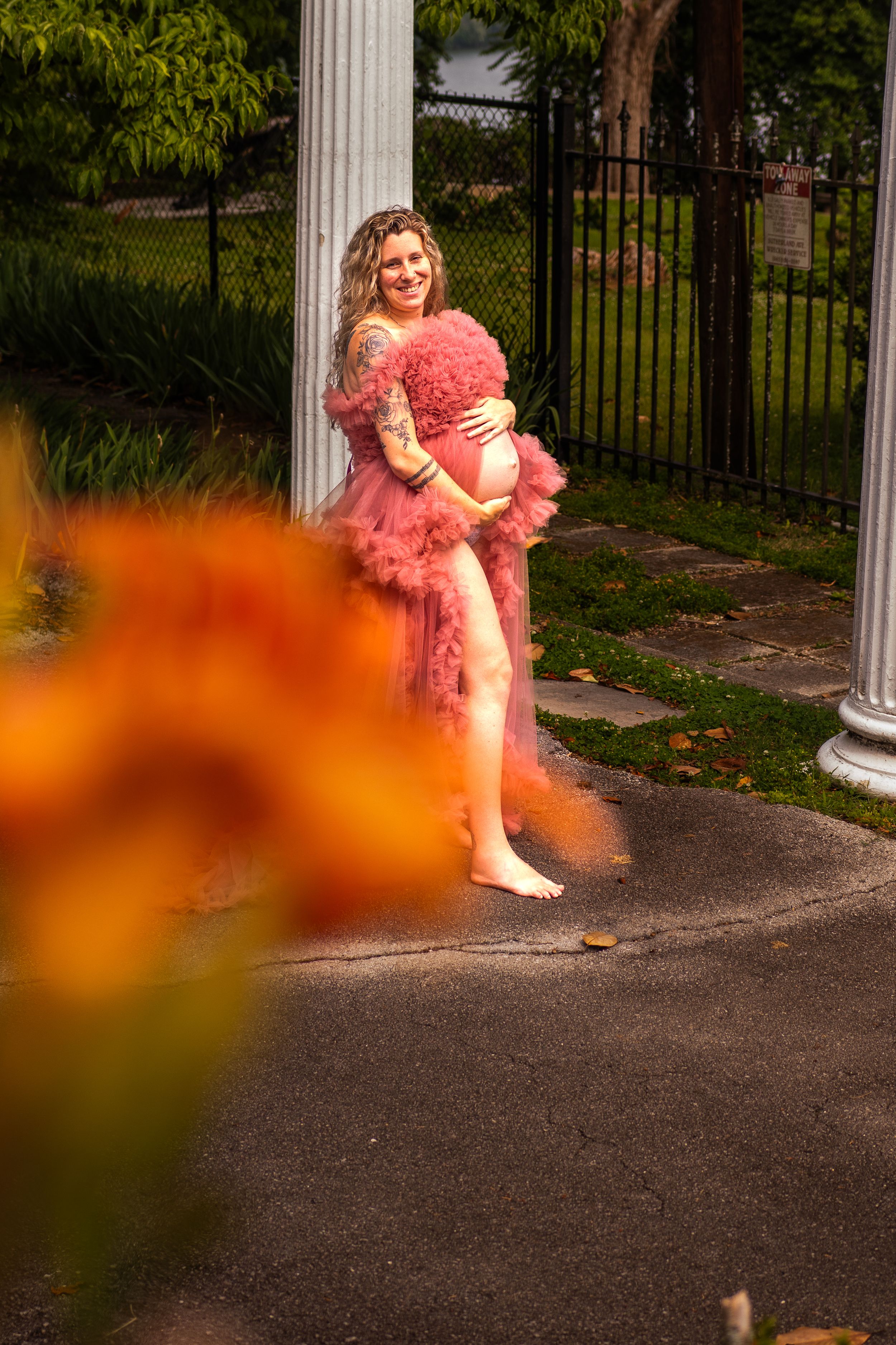 A person in a pink ruffled dress poses next to a white column in a garden setting with orange flowers in the foreground.