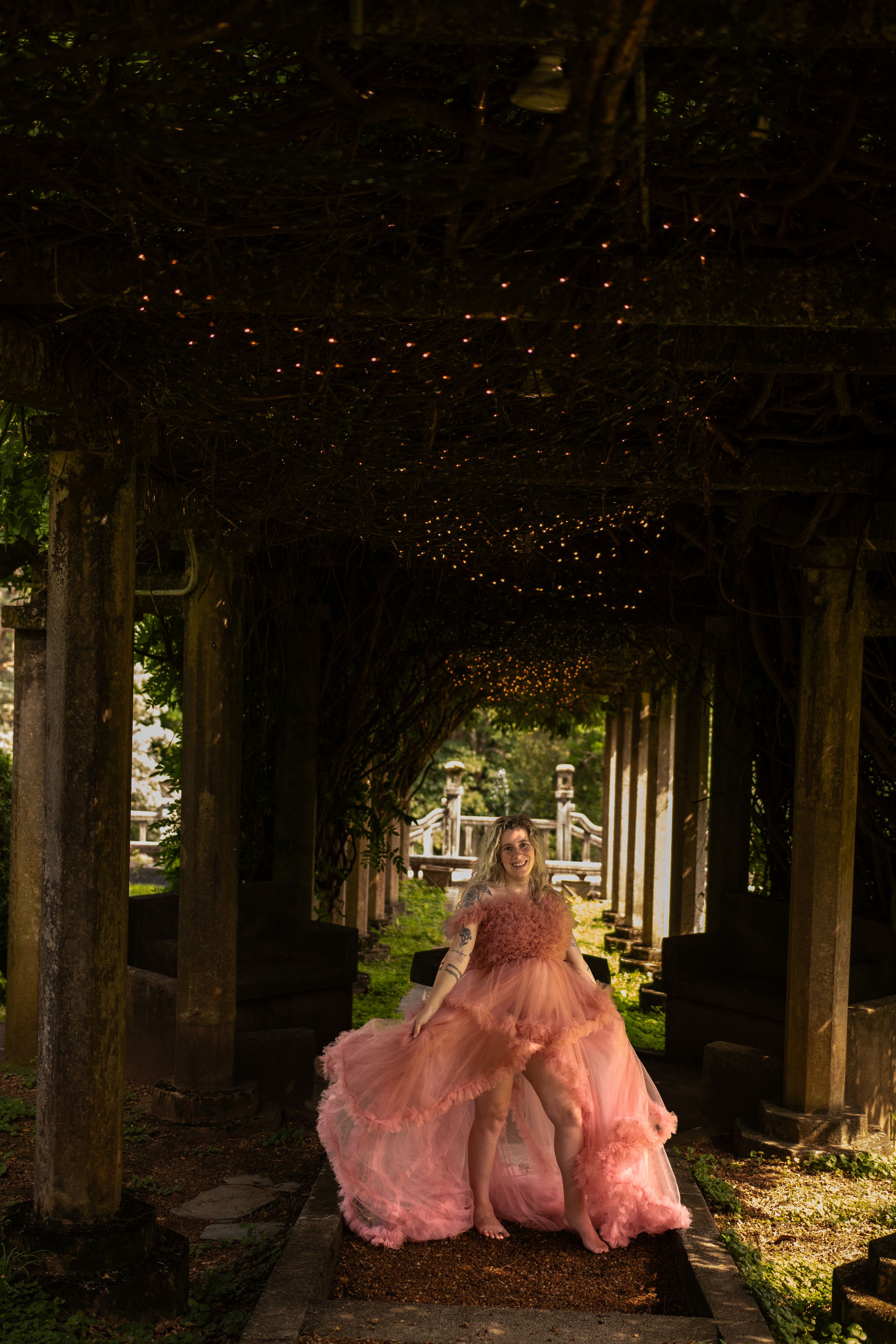 Three pink ballgowns glowing ethereally in a dark wooded colonnade with twinkling lights.