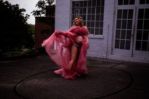 A flowing pink chiffon gown billows dramatically against a white house exterior at dusk.