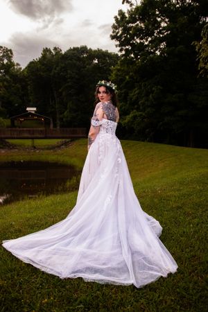 A flowing white wedding dress with a long cape train spreads elegantly across a grassy hillside at sunset.