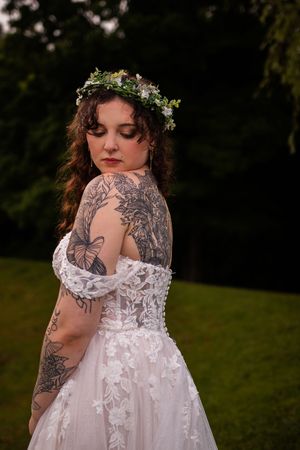 A bride in a lace wedding dress and floral crown shows intricate tattoo artwork on her shoulder and arm.