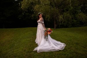 A bride in a flowing white gown and veil poses gracefully on a lush green hillside at sunset.