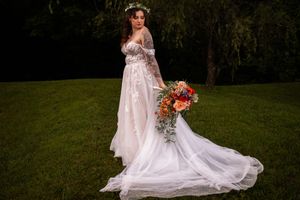 A bride in a flowing white lace gown with off-shoulder sleeves poses in a lush garden setting at sunset.