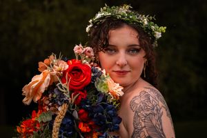 A person wearing a floral crown holds a vibrant bouquet with red and blue flowers against a dark background.