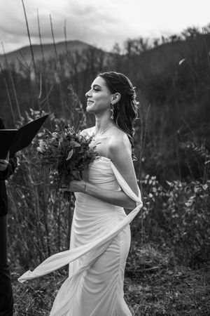 Black and white photo of a bride in a flowing white dress holding a bouquet against a mountain landscape.