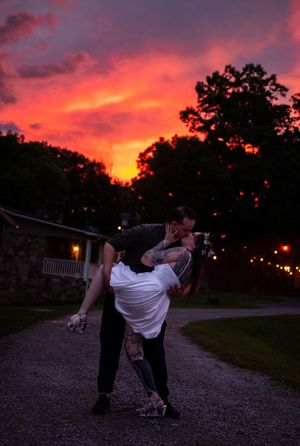 A couple dances on a gravel path at sunset with a dramatic pink and orange sky and string lights glowing in the background.