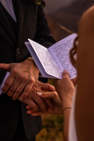 A couple holds hands during a wedding ceremony while reading handwritten vows from a small white book.