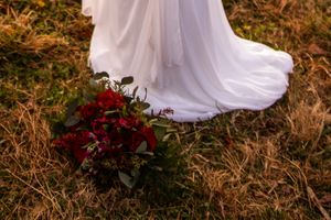 A flowing white wedding dress train with burgundy flowers trails across golden grass in autumn sunlight.