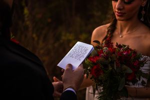 A couple at a wedding ceremony reads from ceremony programs while holding red rose bouquets.