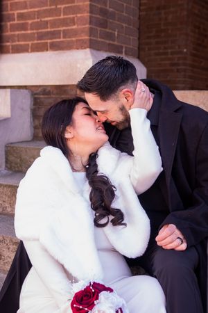 Bride in white fur coat and groom in black suit sharing romantic moment on brick church steps.