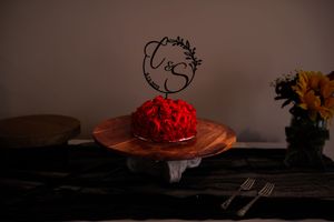 A red cake with a decorative wire topper sits on a wooden pedestal stand in moody lighting.