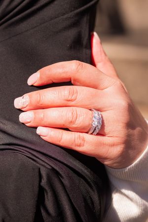 Close-up of a hand wearing a sparkling engagement ring with a white manicure against a black fabric background.