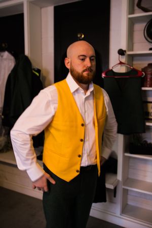 A person wearing a yellow vest and white dress shirt adjusts their clothing while standing in front of mirrors.