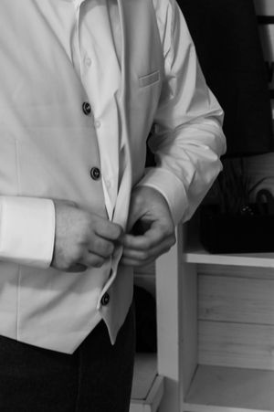 A person wearing a white suit jacket buttons up their formal attire in a black and white photograph.