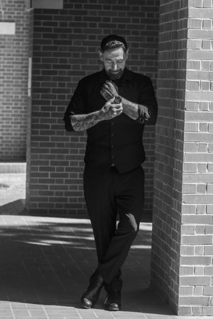 Black and white portrait of person in dark clothing leaning against brick wall while checking phone in sunlit hallway.