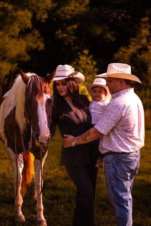 A family stands with their paint horse in a scenic outdoor setting during golden hour, wearing western attire.