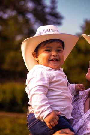 A small child wearing a white hat and shirt smiles while being held outdoors during golden hour.