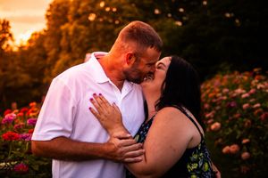 Romantic couple embraces in flower garden during golden sunset with pink and orange sky.