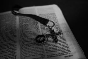 A rosary with black beads and cross rests on an open Bible page in dramatic black and white lighting.