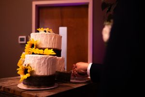 White wedding cake decorated with sunflowers sits on a dark table under warm lighting.
