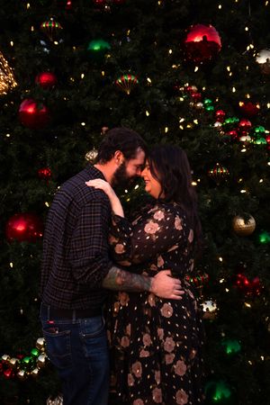 A couple shares an intimate moment in front of a decorated Christmas tree with twinkling lights and ornaments.